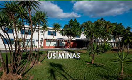 Fachada do Centro de Memória Usiminas cercada por palmeiras e um gramado verde, sob um céu claro.