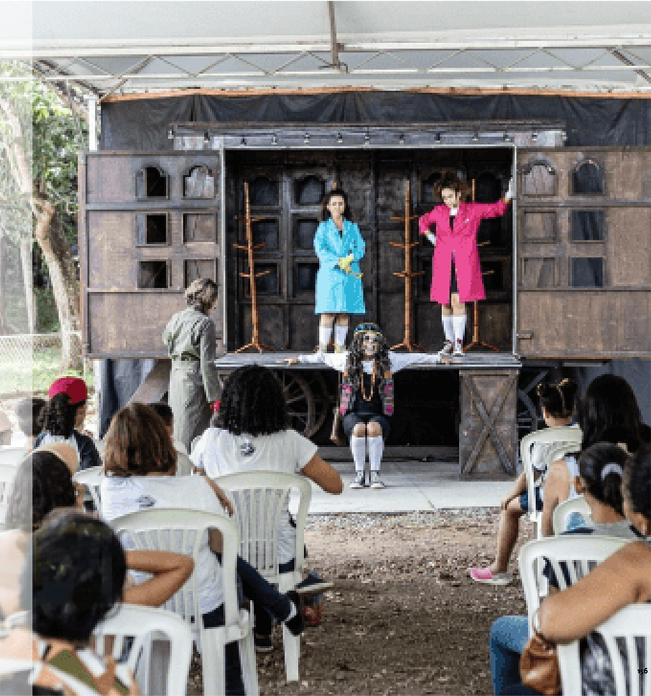 Cena de uma peça teatral ao ar livre, com três atores vestidos com trajes coloridos se apresentando em um palco elevado, enquanto um público diversificado de adultos e crianças assiste sentado em cadeiras brancas de plástico.
