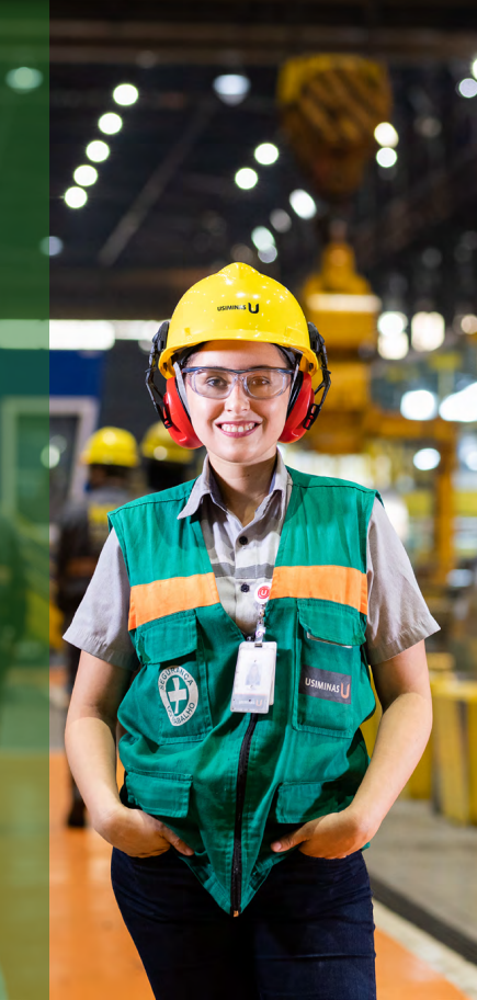 Funcionária da Usiminas sorridente, vestindo equipamento de segurança completo, incluindo capacete, óculos, protetores auriculares e colete refletivo, em ambiente industrial.