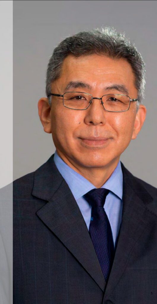 Homem asiático de meia-idade, com cabelos grisalhos curtos e óculos, vestindo um terno escuro, camisa azul clara e gravata azul escura, olhando diretamente para a câmera e sorrindo levemente.